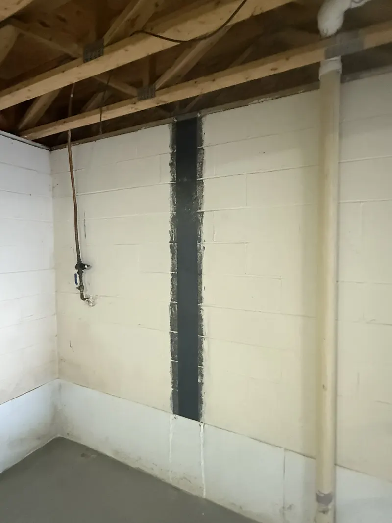 A single vertical carbon fiber strap on a white block basement wall below exposed floor joists