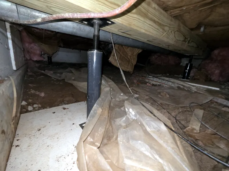 Adjustable steel support jack with a threaded top and plate under a wood beam, with plastic sheeting on the crawl space floor