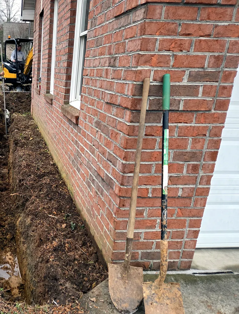 Drainage trench dug along a brick wall, with two shovels and a mini excavator working behind