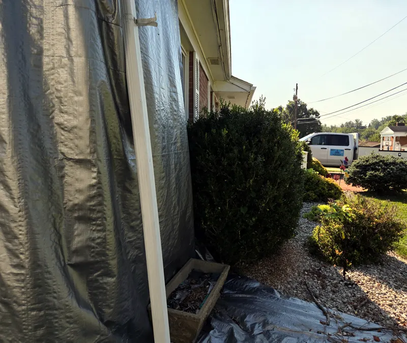 Side of a house wrapped in tarps during exterior work, with an MCA Foundations work truck in the driveway