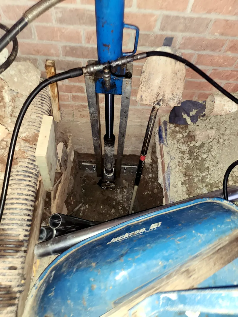 Hydraulic jack and steel pier being installed in an excavation at a brick foundation